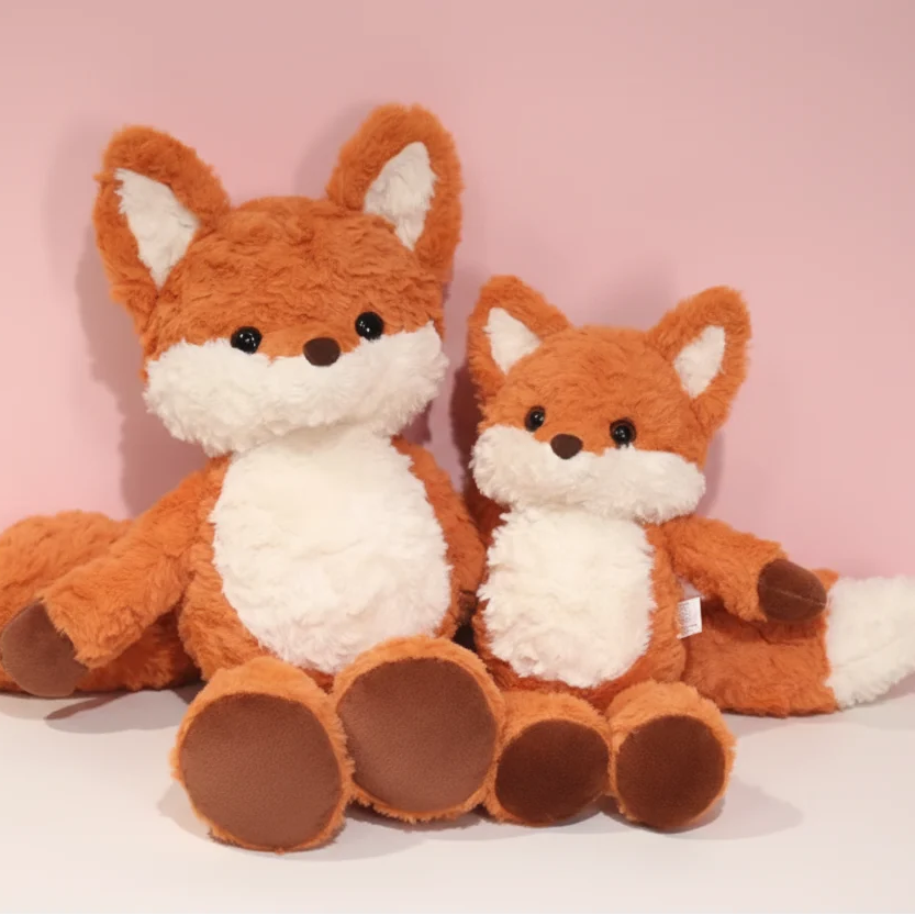 Two plush fox toys in different sizes sitting side by side against a soft pink background.