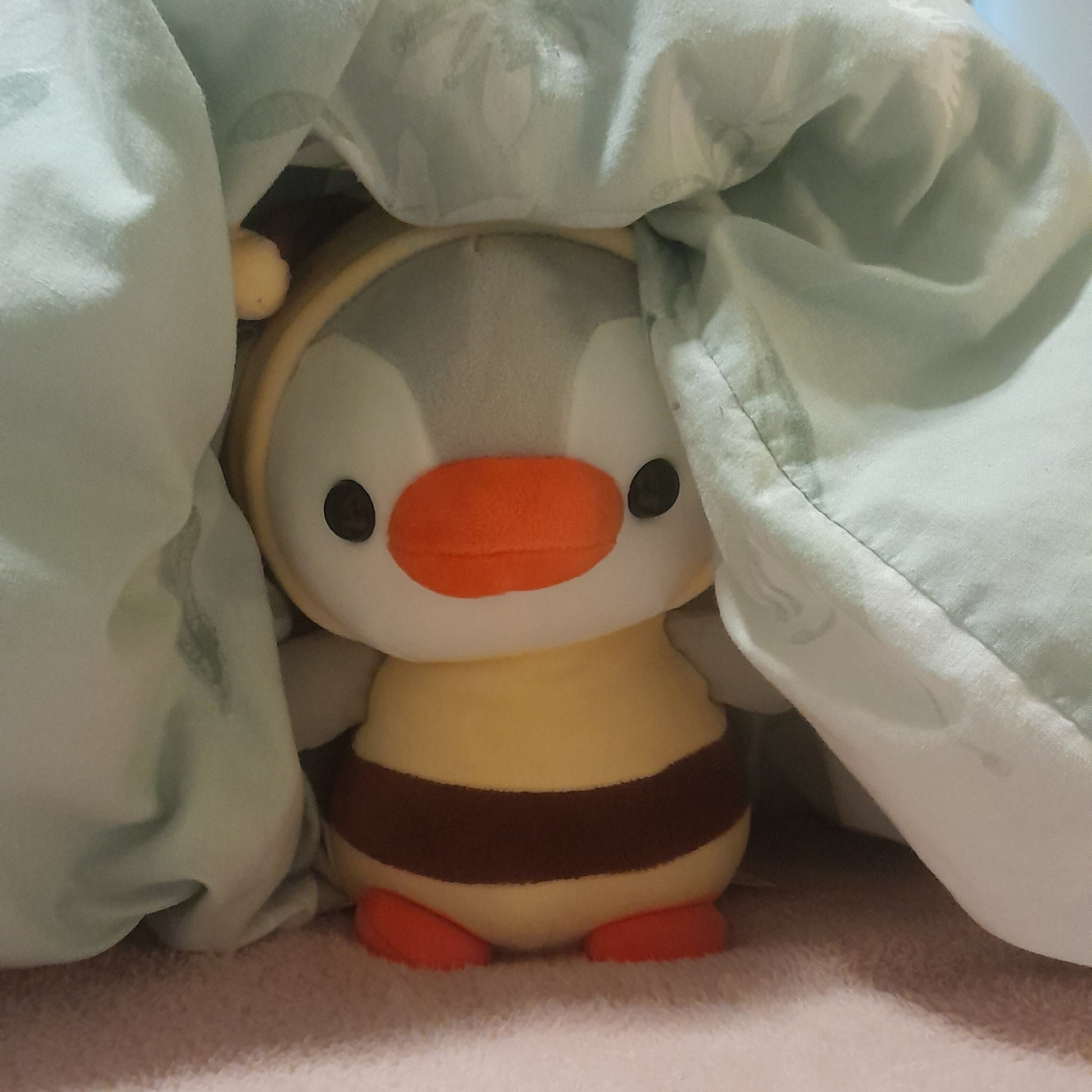 A small plush toy penguin wearing a yellow and brown bee outfit peeks out from under a light mint green blanket on a pink surface.
