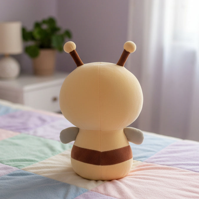 A plush toy shaped like a cartoon bee or insect, shown from the back on a white surface against a gray wall. The toy is cream-colored with dark brown antennae and a dark brown horizontal band around the middle.