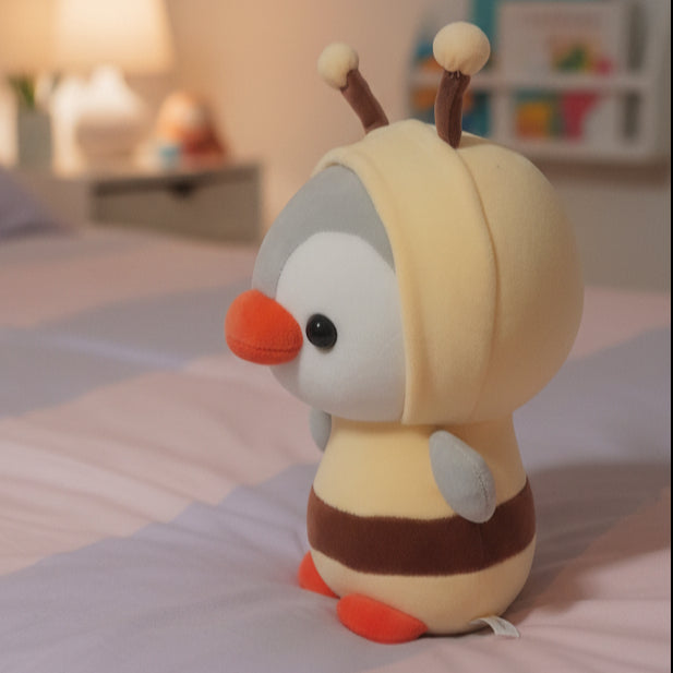 A plush toy character dressed in a bee costume, featuring a cream-colored hood, brown antennae, a gray face, orange beak, and brown and orange striped body, standing next to a small white speaker.
