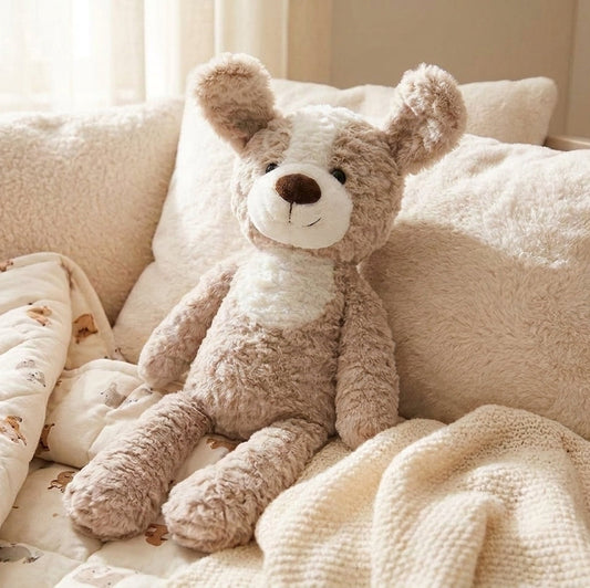 A plush puppy dog toy with brown and cream fur and upright ears, sits on a bed surrounded by cream bedding, pillows, and a blanket in a softly lit bedroom.