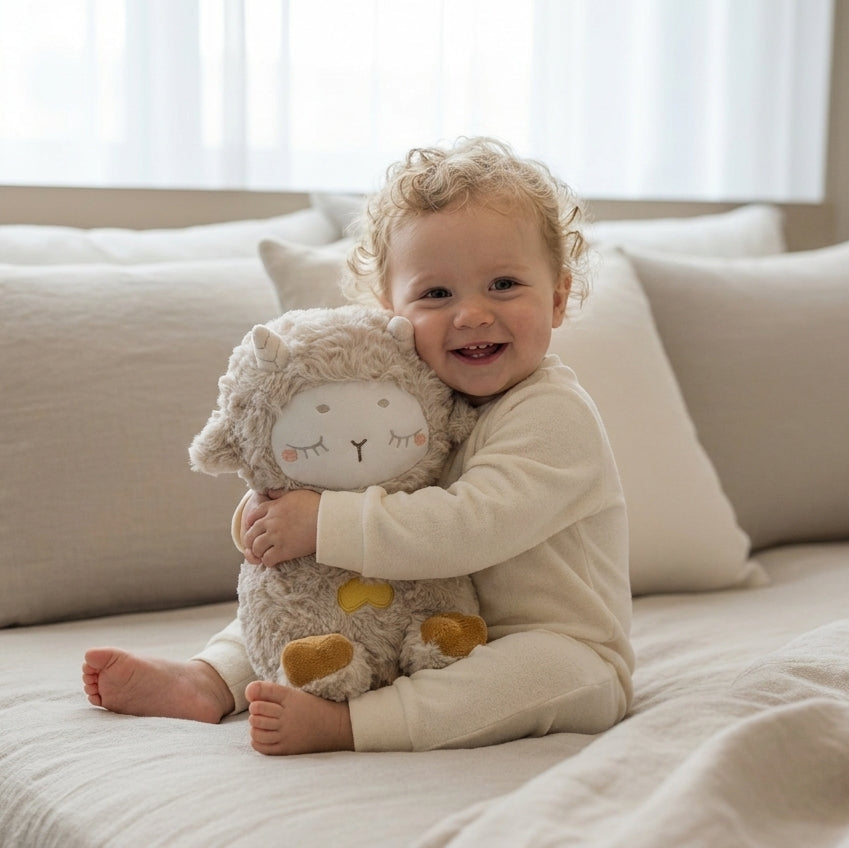 A smiling toddler with curly blonde hair sits on a bed holding a plush sheep toy. The child is wearing cream-colored pajamas and is surrounded by neutral beige pillows and bedding in a softly lit bedroom.