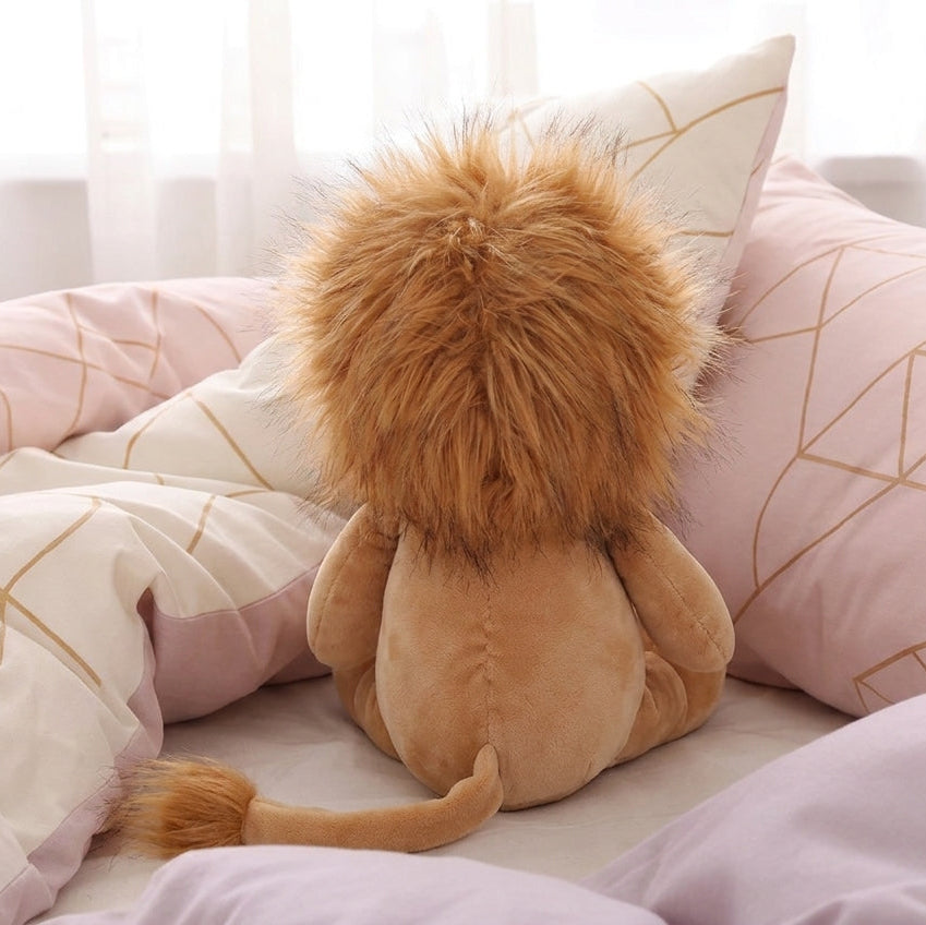 A plush lion toy sitting on a bed with pink and cream bedding, viewed from behind to show its fluffy golden-brown mane and tan body.