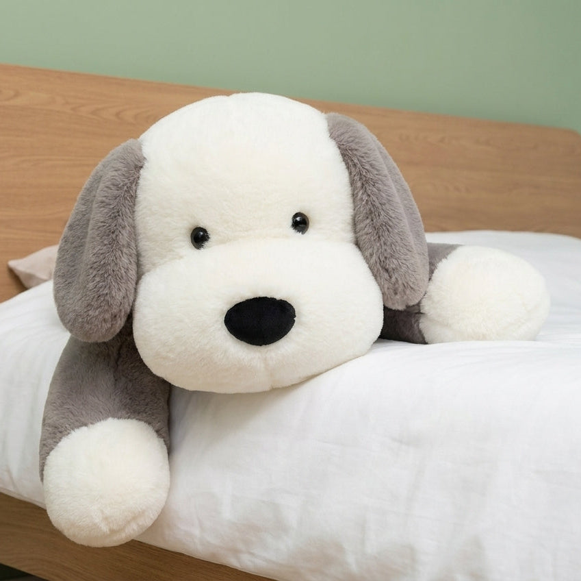 A plush dog toy with a white face and paws, gray floppy ears, and a black nose, resting on a white bed with a wooden headboard against a light green wall.