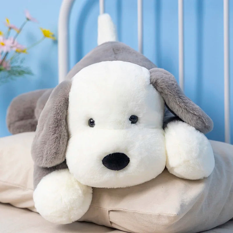 A soft plush toy puppy with gray ears and a beige body, lying on a bed.