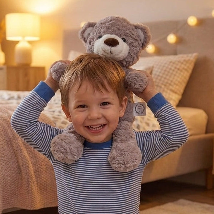 A smiling young child wearing blue and white striped long-sleeve pajamas sits on a carpet in a warmly lit bedroom, holding a gray plush teddy bear above their head.