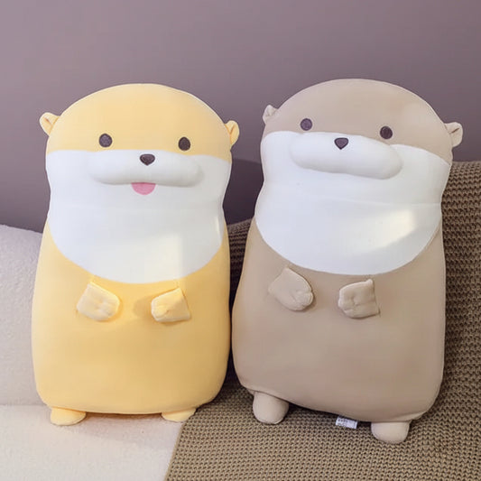 Two soft otter pillow toys, one yellow and one beige, sitting on a sofa.