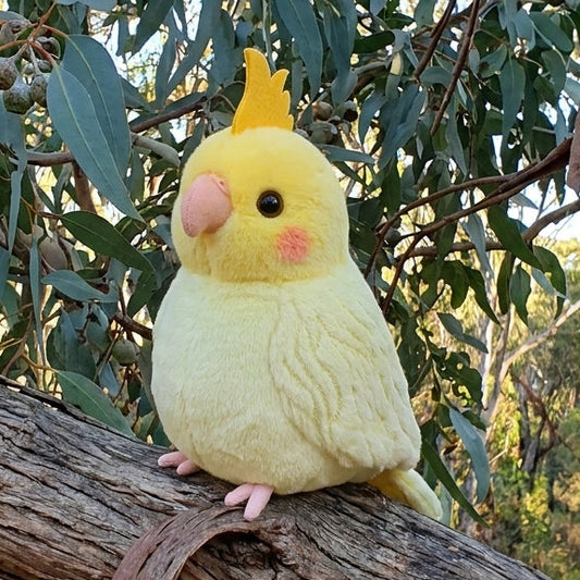 A plush toy cockatiel in pale yellow with a small yellow crest, pink beak, and pink feet, perched on a tree branch outdoors with eucalyptus leaves in the background.