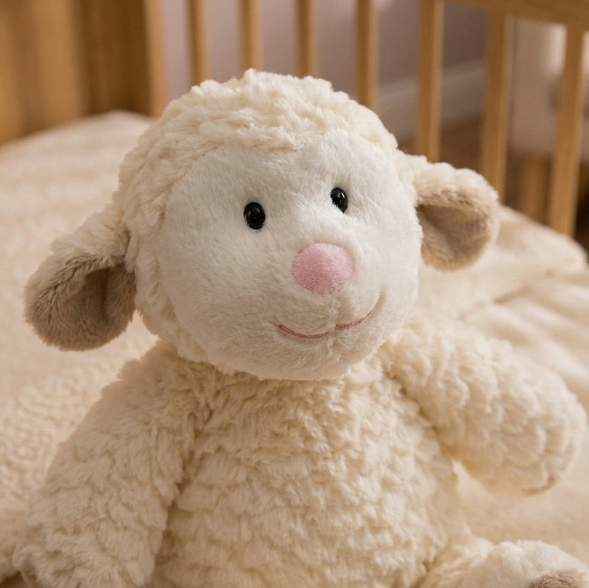 A plush cream-colored stuffed sheep toy with a pink nose and black eyes, sitting on a soft cream blanket inside a wooden crib.