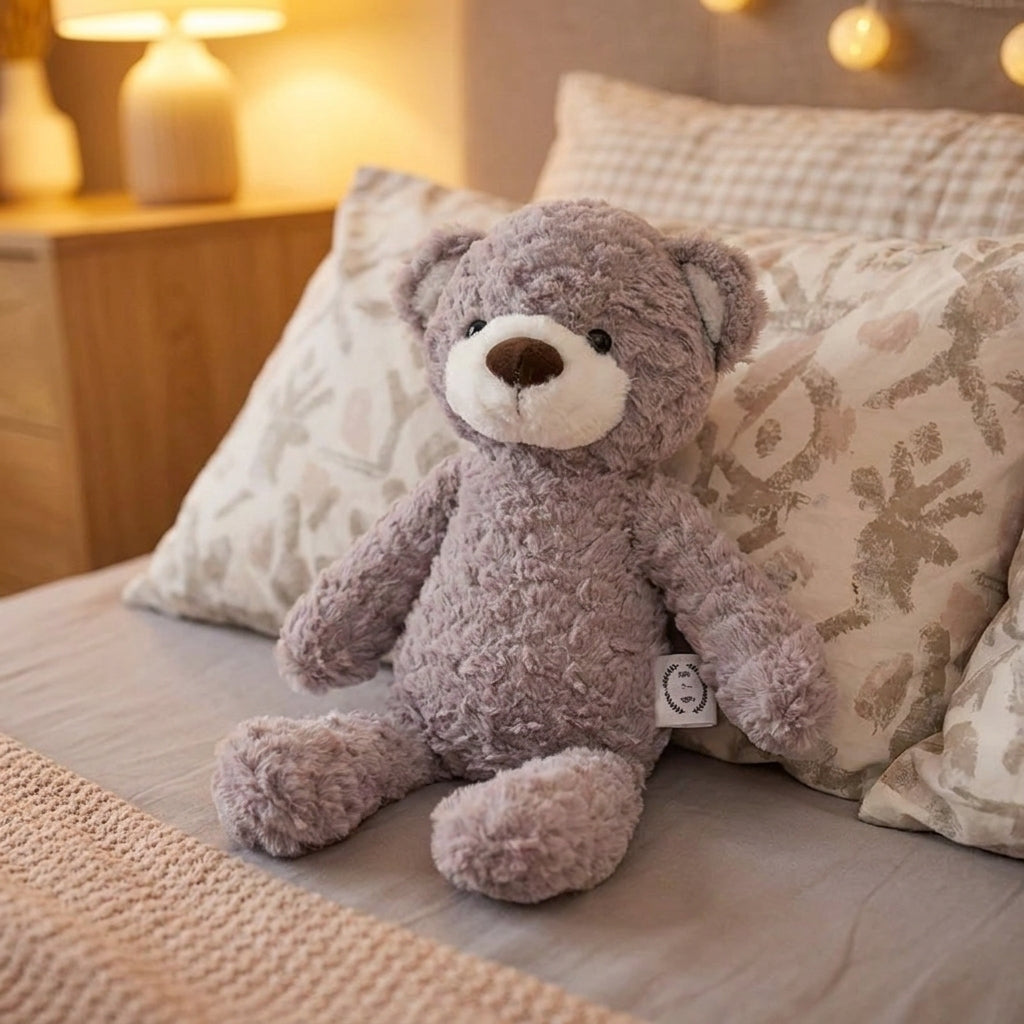 A plush teddy bear in a dusty lavender-gray color sits on a bed with patterned pillows and a warm bedside lamp in the background.