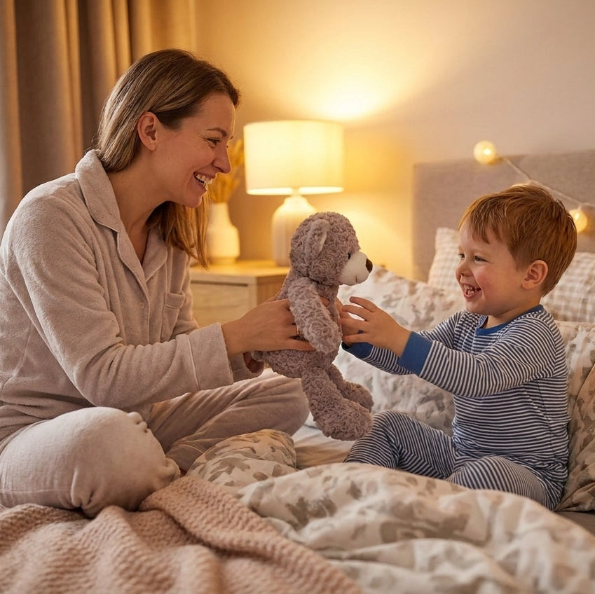 A woman and a young boy sitting on a bed in a warmly lit bedroom, smiling and playing with a gray stuffed teddy bear. The woman wears light beige pajamas, and the boy wears blue-and-white striped pajamas.