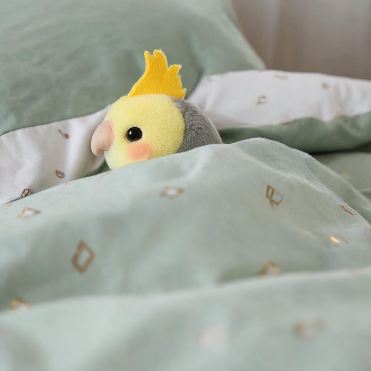 A small plush bird toy with a yellow crest and gray body peeking out from under a mint green patterned duvet on a bed.